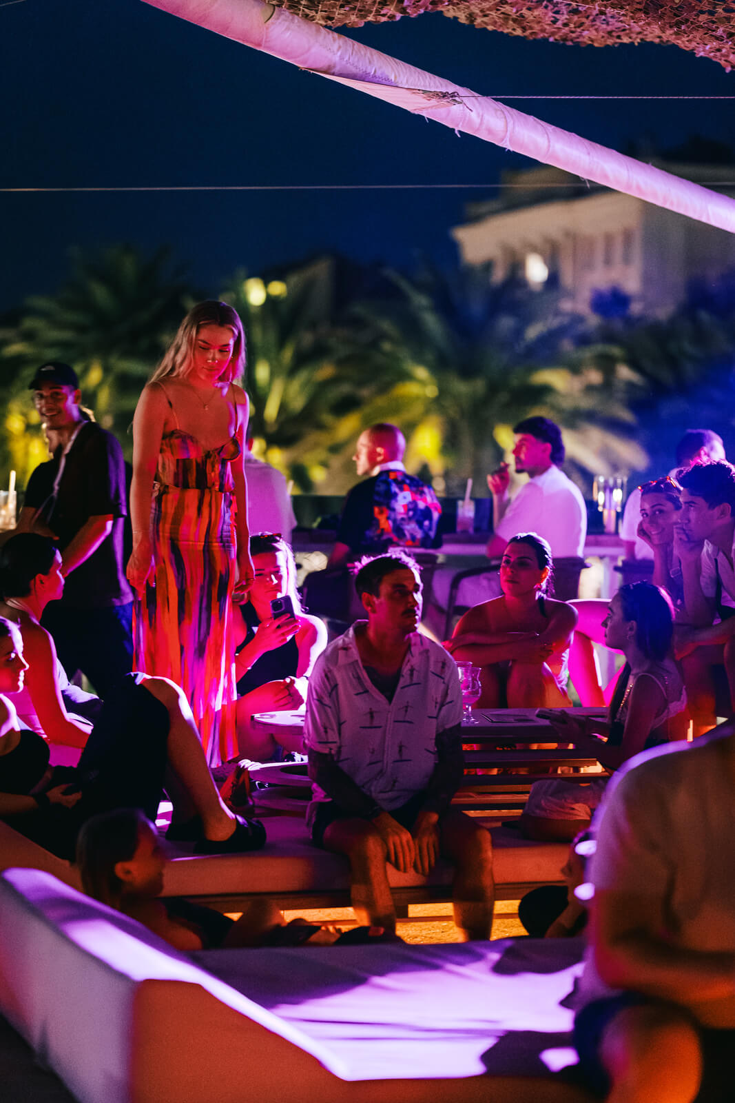 People relaxing and socializing on lounge beds and low seating on a rooftop at night, illuminated by dramatic pink and purple mood lighting. Palm trees are visible in the background.