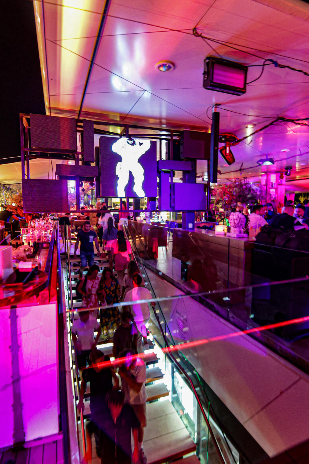 Nightlife scene with colorful lighting, bar area, and people walking down illuminated stairs.