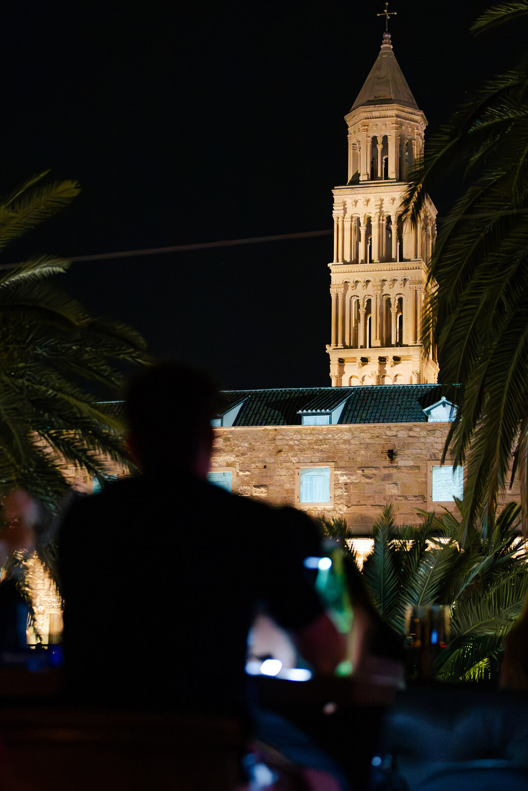 Silhouette of a person sitting at an outdoor bar at night with a lit historic tower in the background.