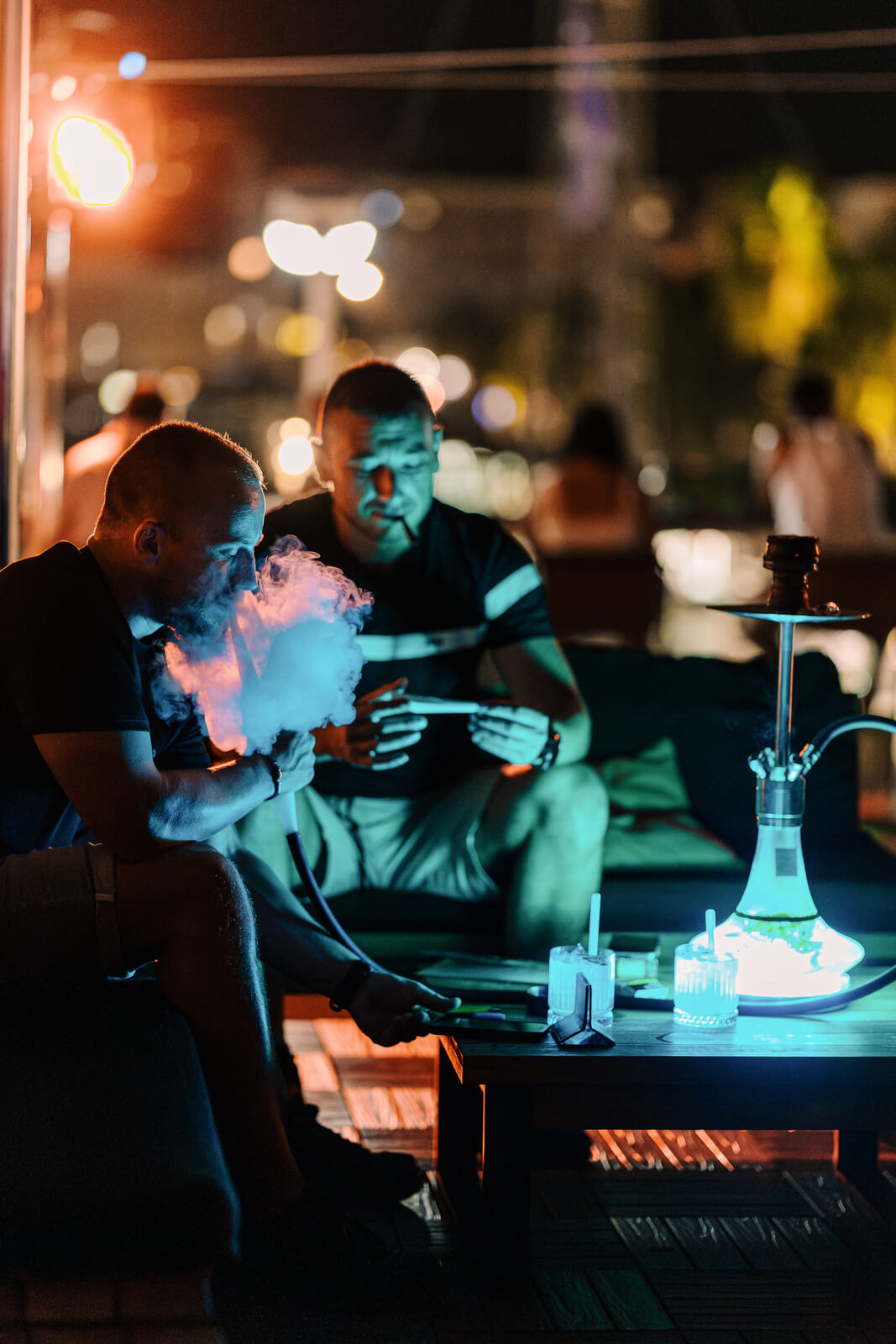 Two men relaxing on a dimly lit rooftop patio at night, with one exhaling a large cloud of glowing blue smoke from a hookah pipe. A colorful, illuminated hookah sits on a table next to them.