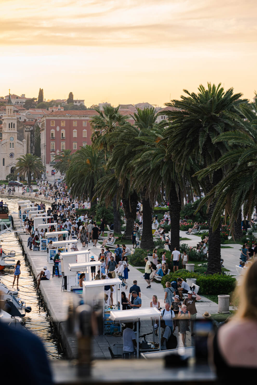 Busy waterfront promenade lined with palm trees and people walking along the marina at sunset.