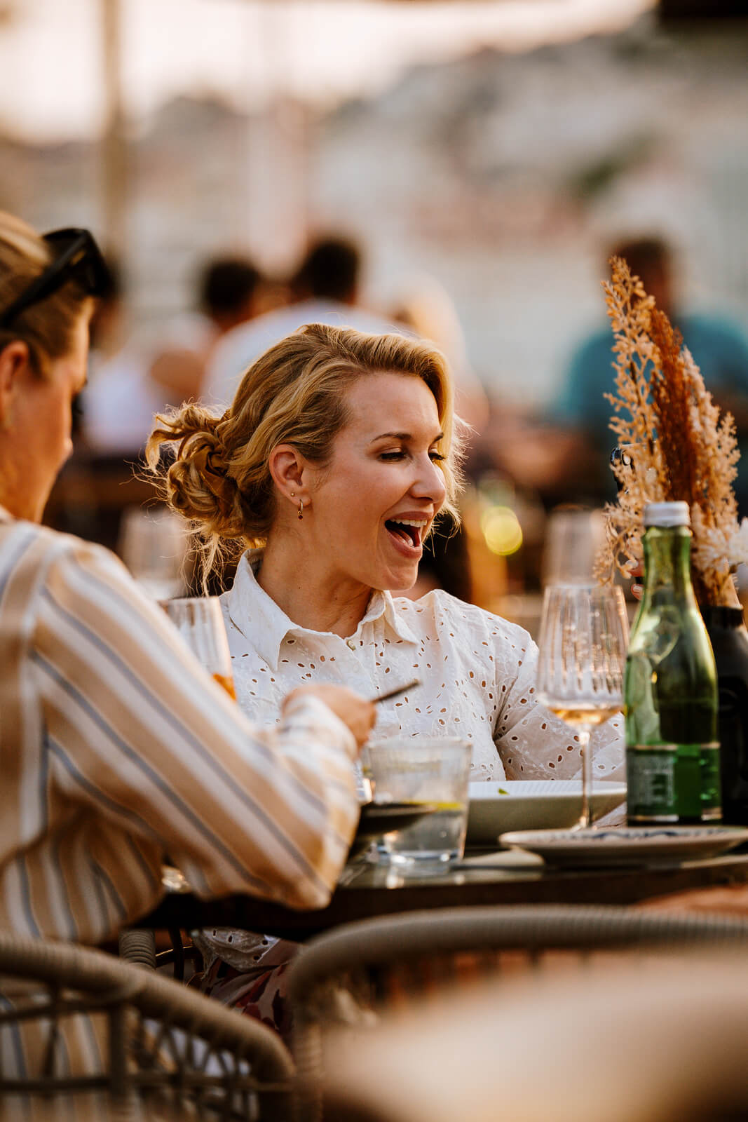 Woman laughing while dining outdoors at sunset, surrounded by glasses and decor.