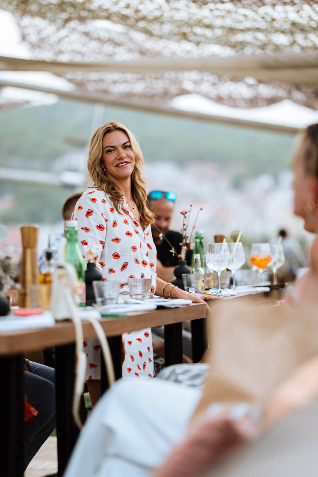 A smiling blonde woman in a white dress with red lip prints standing next to a table at a brightly lit outdoor dining area. Various drinks and glassware are visible on the wooden table.