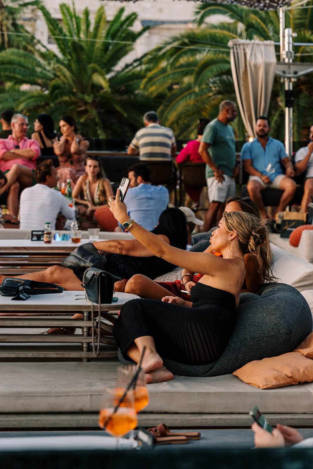 Woman sitting on a lounge chair taking a selfie while people socialize at a rooftop bar during sunset.