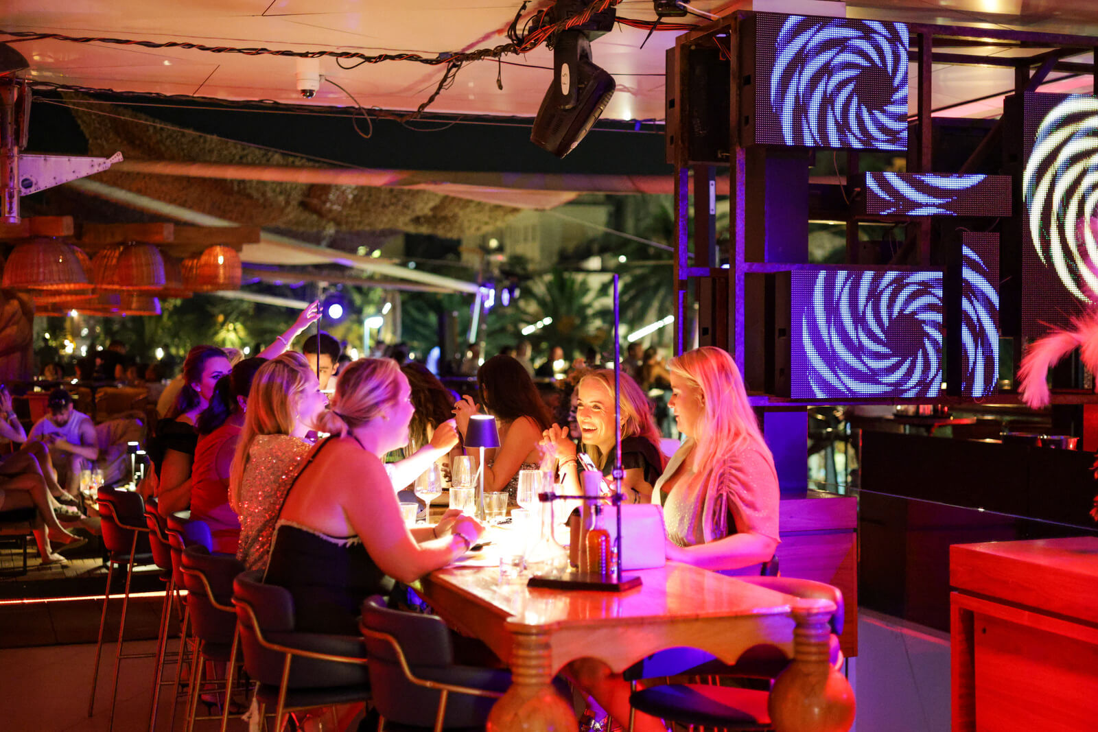 Group of women enjoying drinks and conversation at a lively bar with neon lighting.