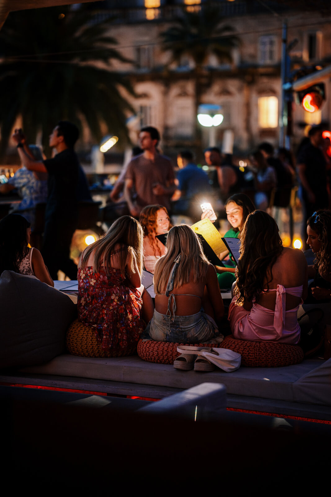 Group of women sitting on lounge cushions, illuminated by phone light while reading menus at night.