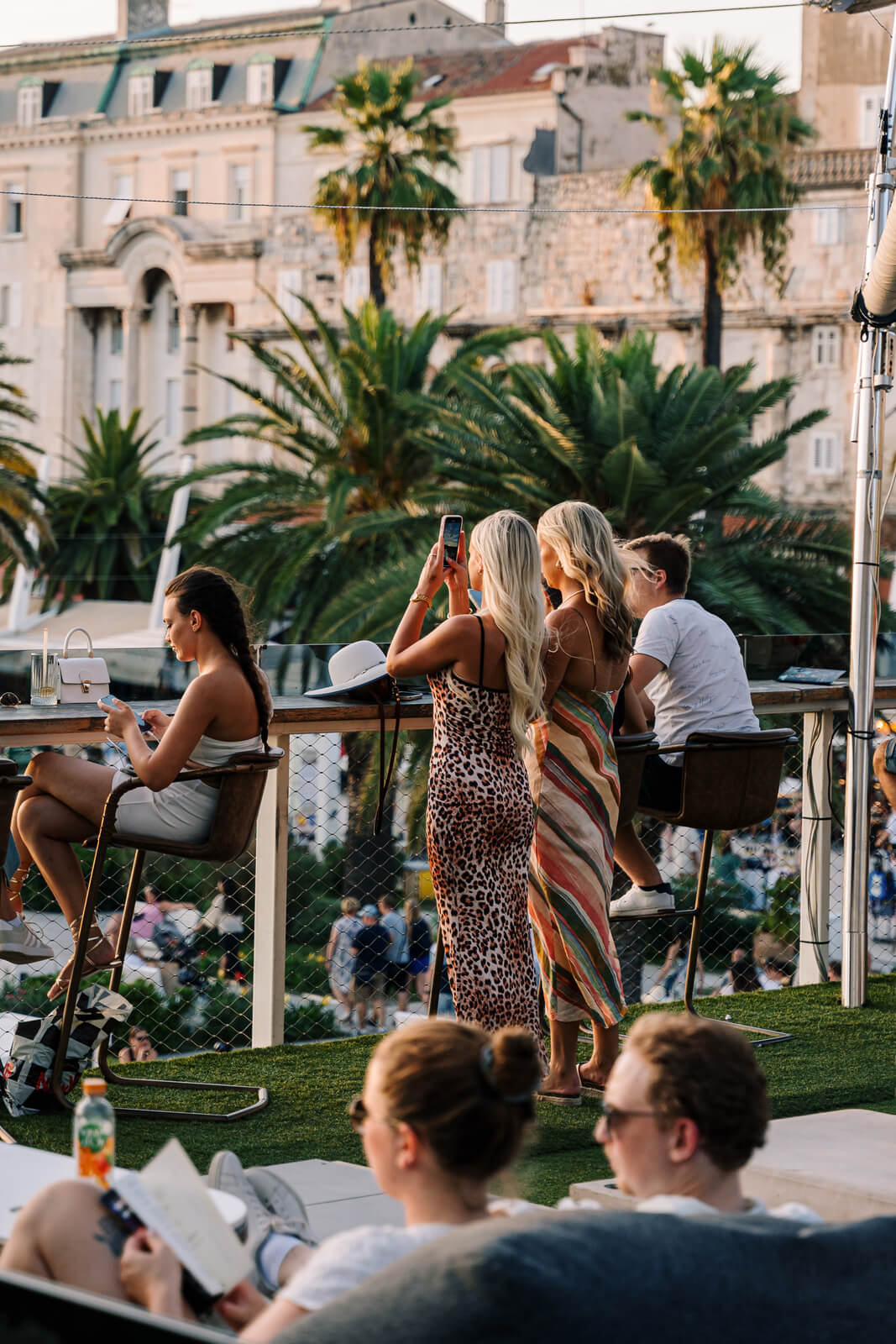 Women standing and taking photos at a rooftop bar with palm trees and historic buildings in the background.