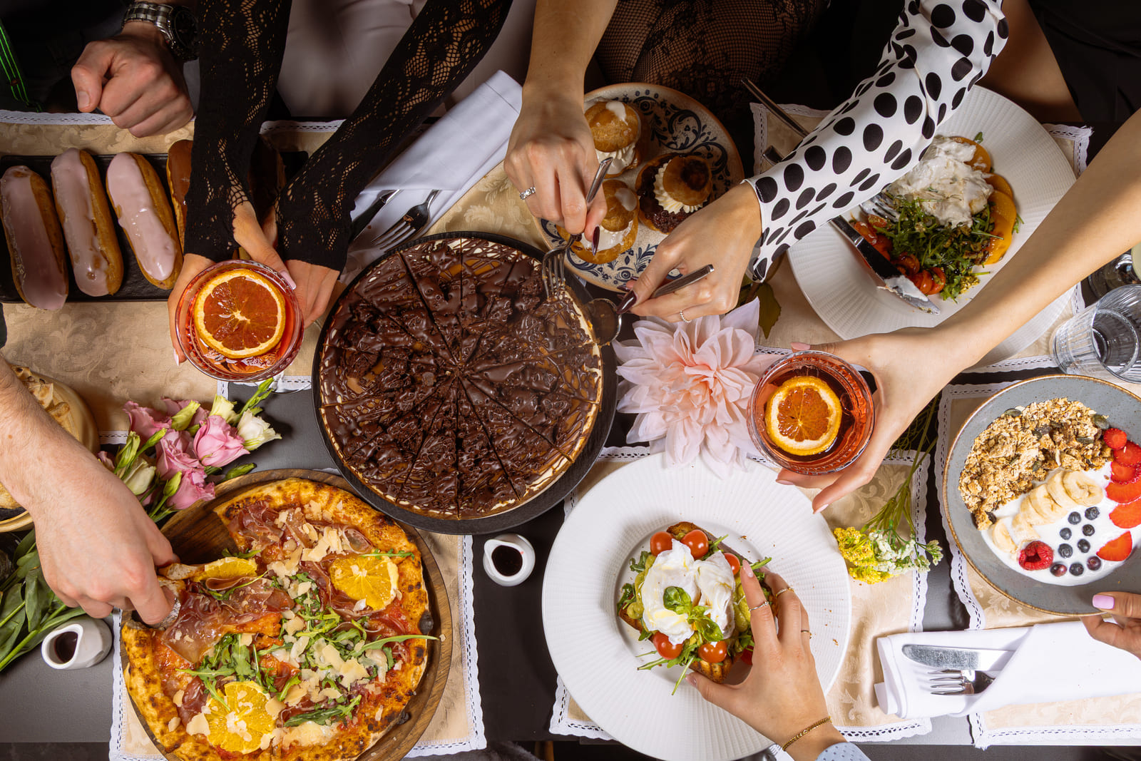 A vibrant brunch spread viewed from above, featuring chocolate cake, éclairs, fresh pizza with orange slices, burrata salad, granola bowls, and people’s hands reaching for food and drinks.