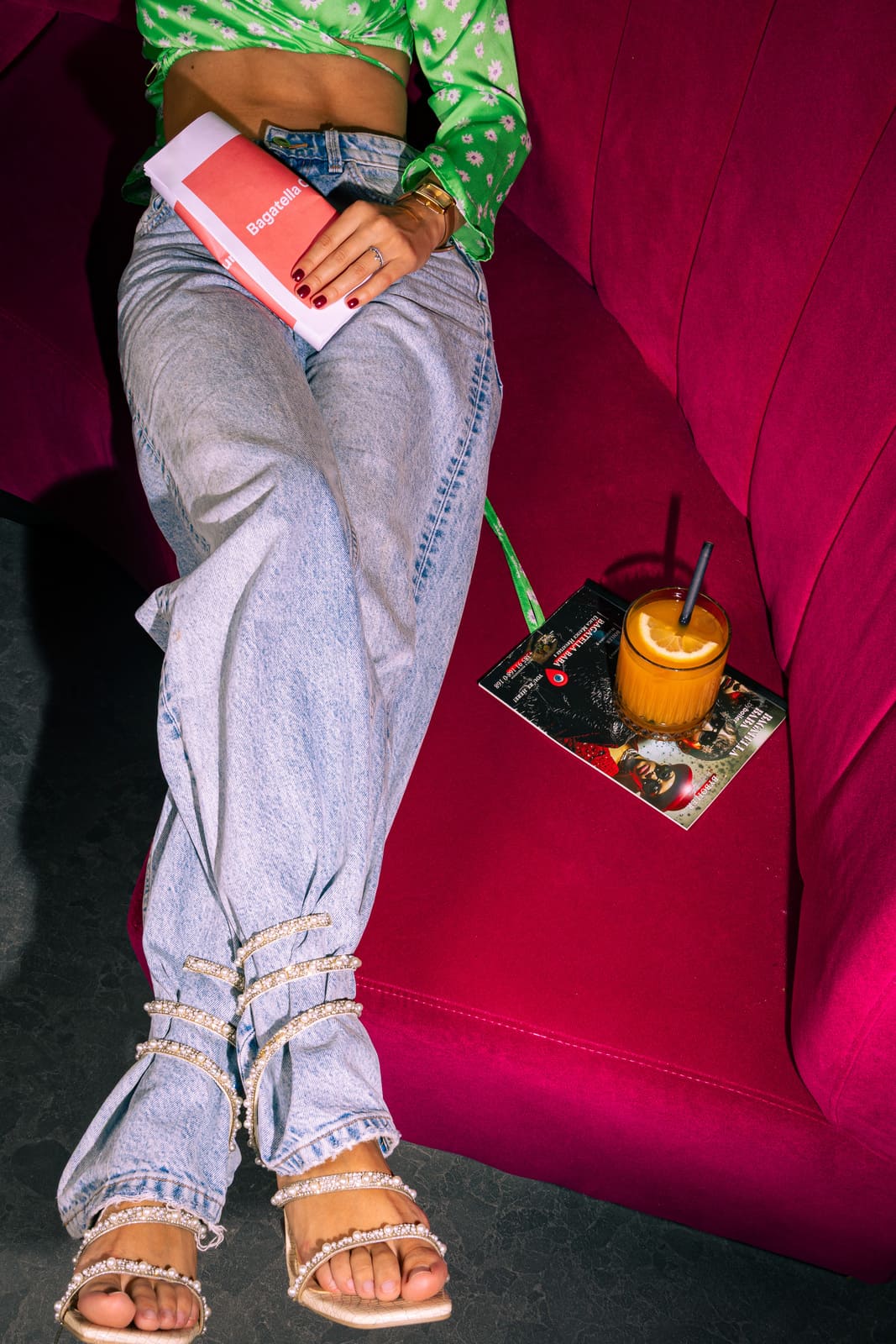 A woman lounges on a magenta couch wearing light blue jeans and crystal-embellished heels, holding a folded menu labeled “Bagatella” with a citrus cocktail resting beside her.