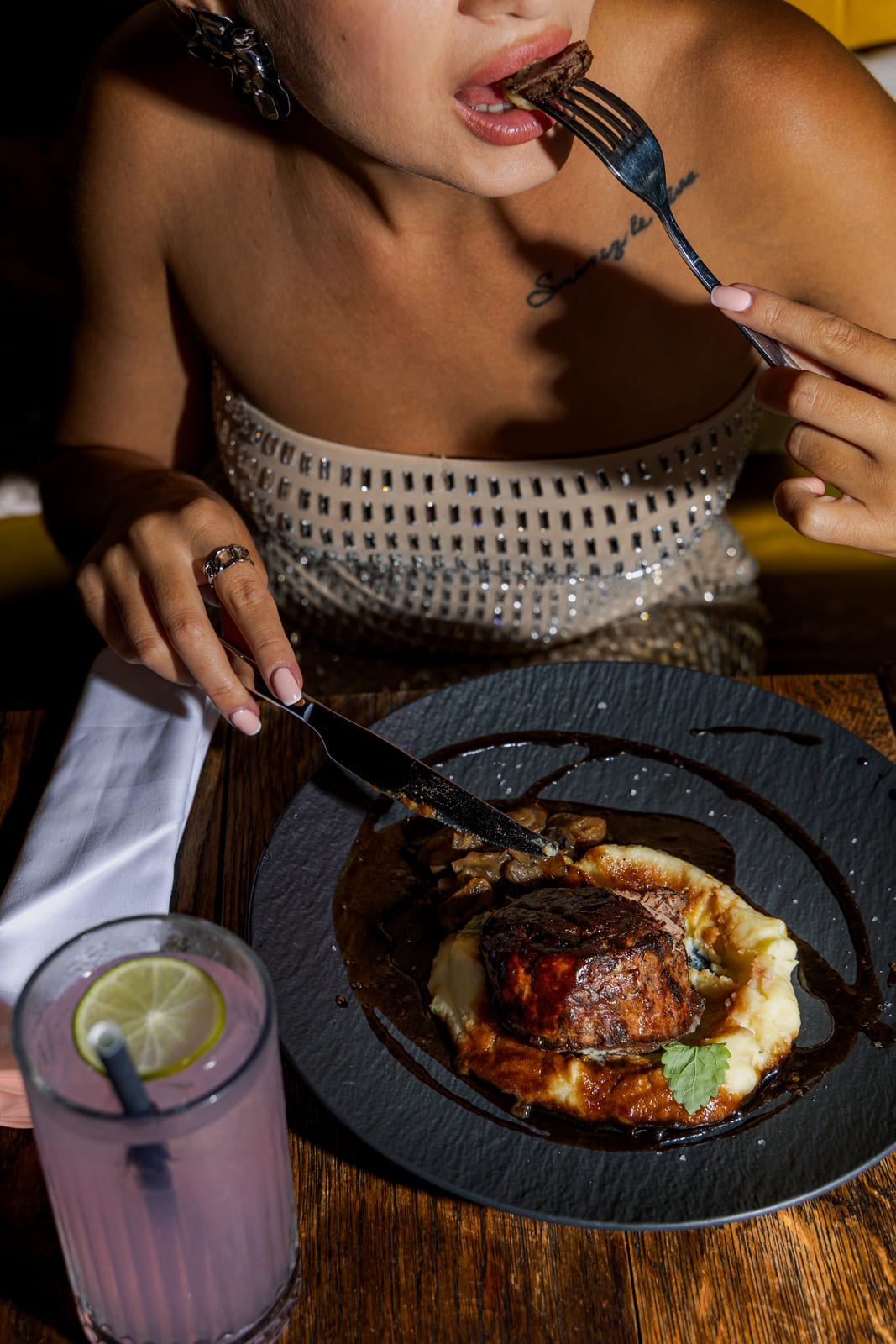 A woman in a rhinestone-studded strapless dress takes a bite of a steak dish, which is plated with mashed potatoes, sautéed mushrooms, and a dark glaze on a black plate.