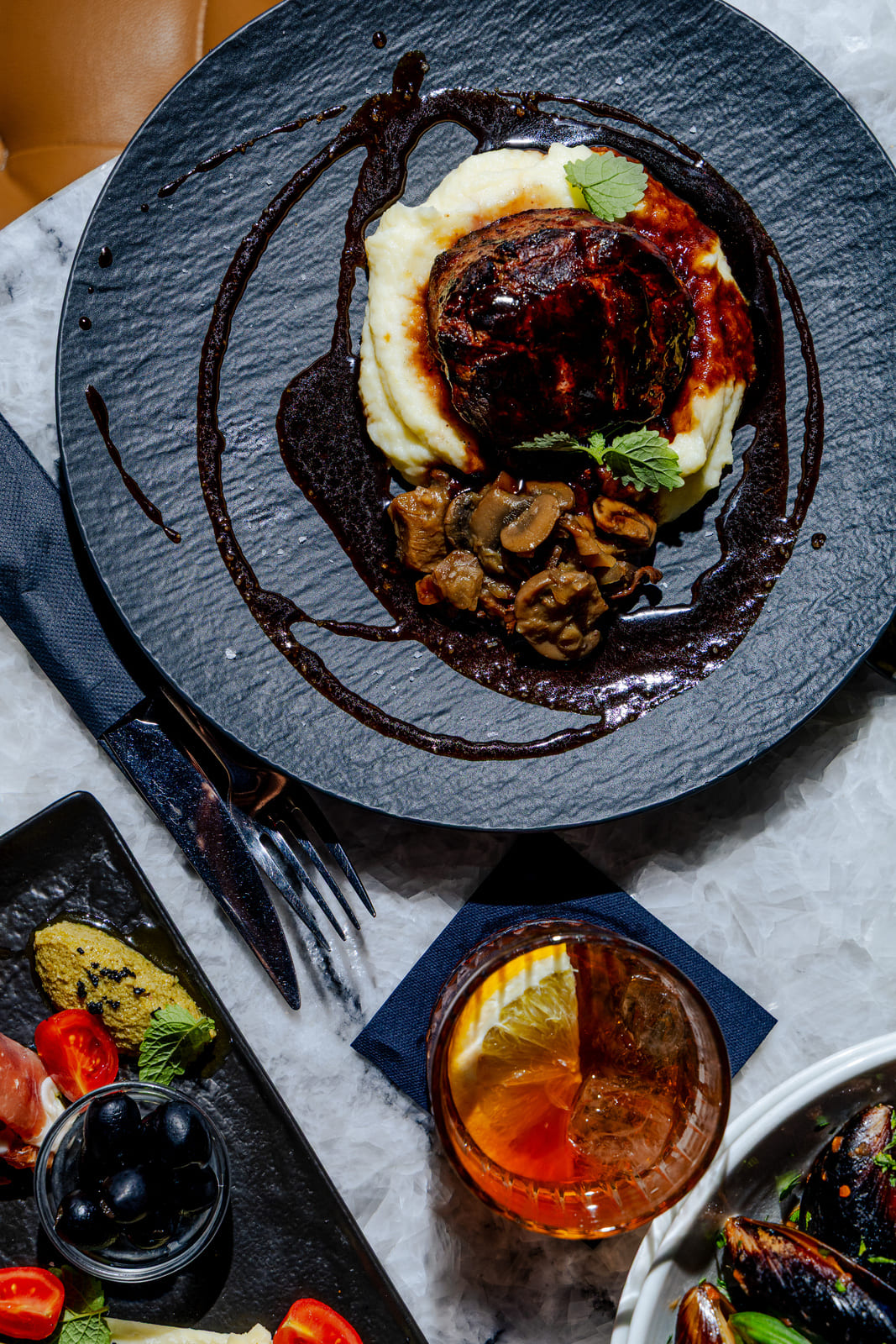 Close-up of a steak dish on a black plate with creamy mashed potatoes, sautéed mushrooms, and a balsamic glaze, paired with an orange cocktail on a white marble table.