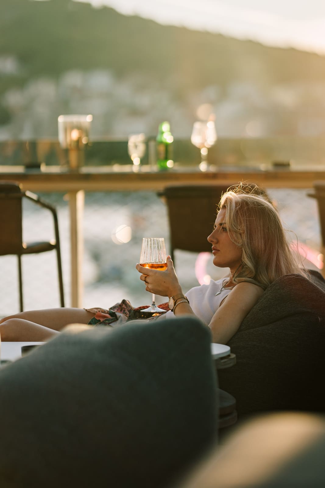 A woman relaxes with a glass of wine at an outdoor rooftop lounge during golden hour, gazing out toward the waterfront with a view of mountains in the background.