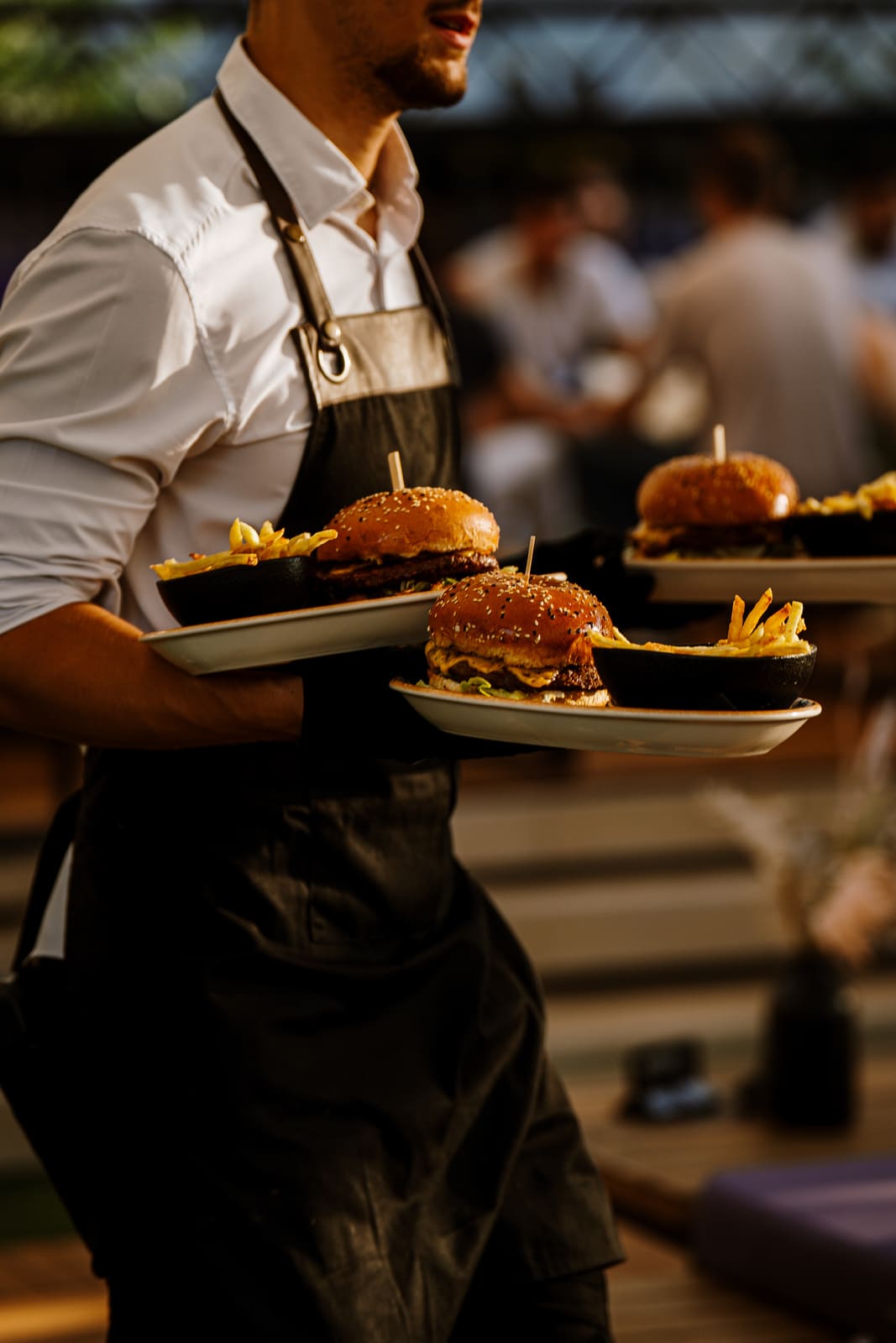 Waiter wearing a white shirt and black apron serving three plates with gourmet cheeseburgers and French fries at an outdoor dining area.