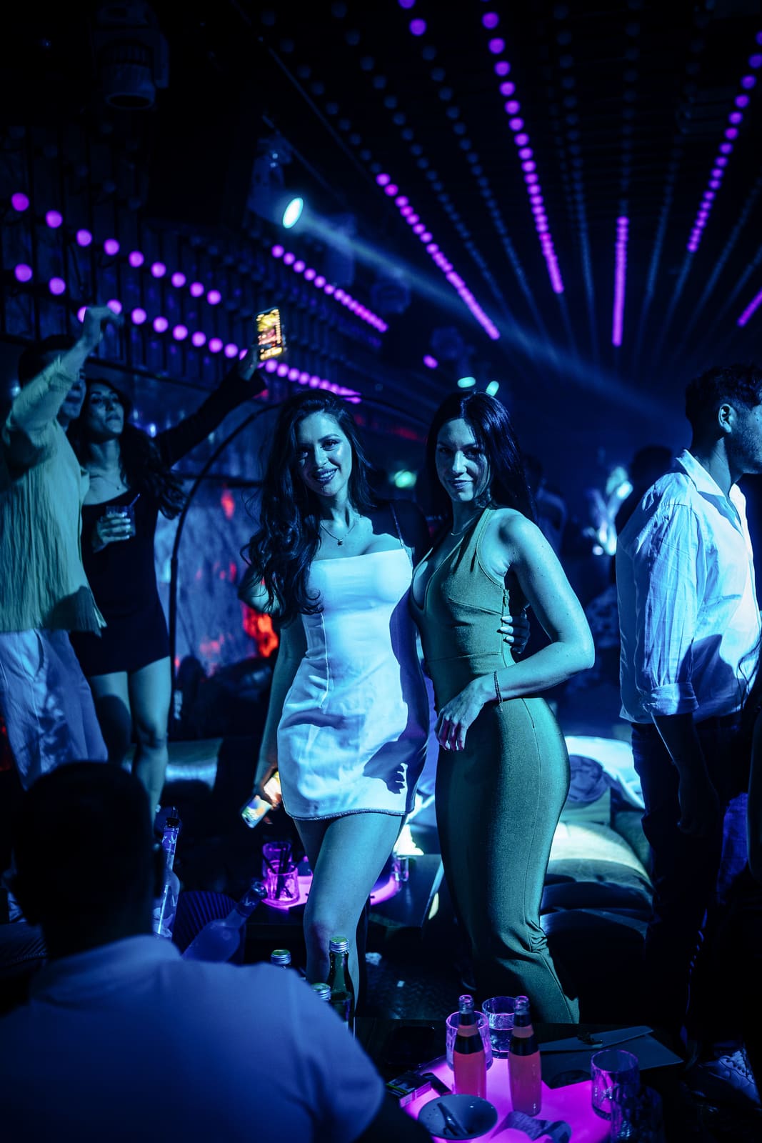 Two women posing together under purple and blue club lights, surrounded by a lively crowd, with LED ceiling lights creating a vibrant tunnel effect.
