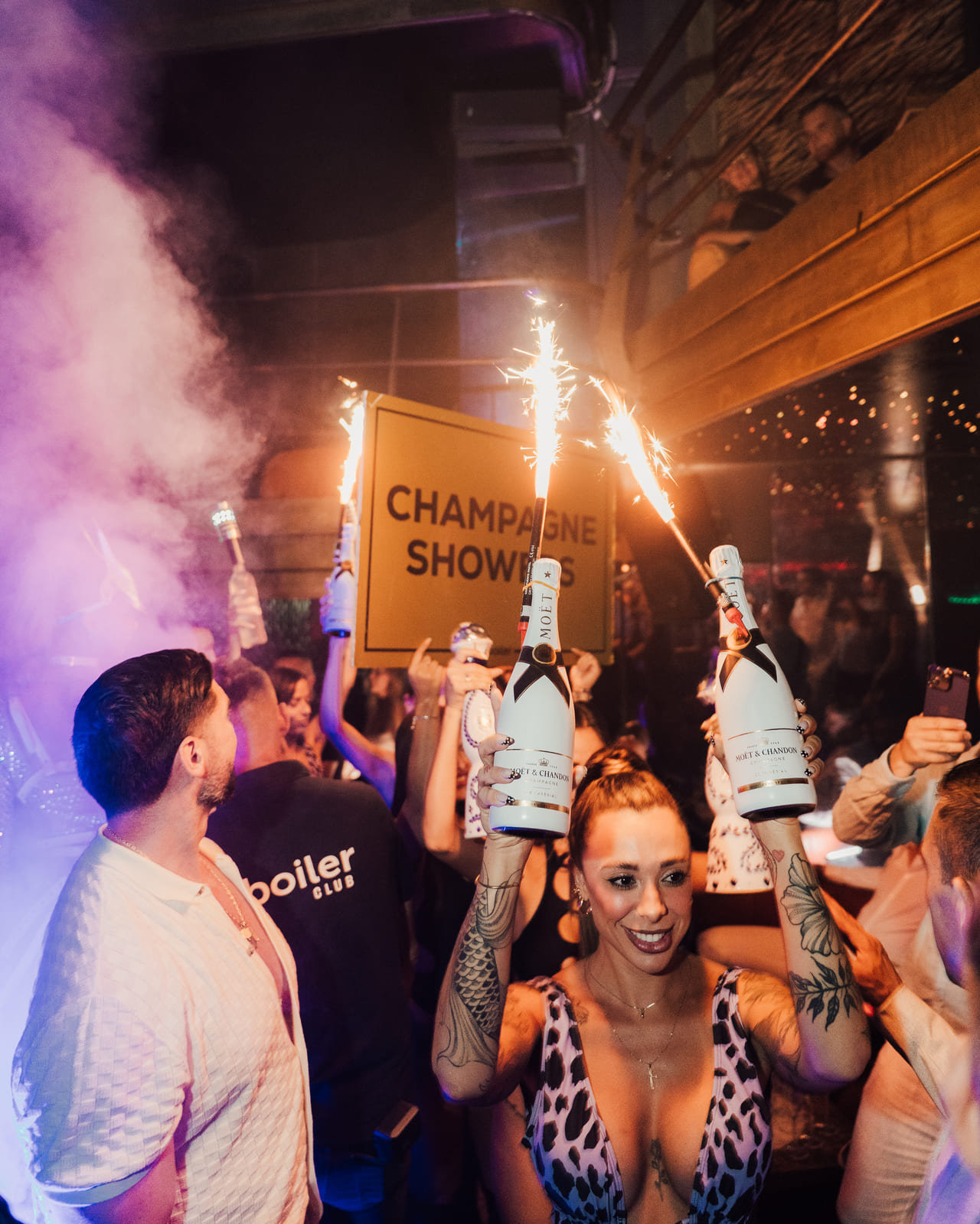 Woman in leopard print top smiling and holding two Moët champagne bottles with sparklers in a crowded nightclub, surrounded by guests celebrating a champagne shower moment.