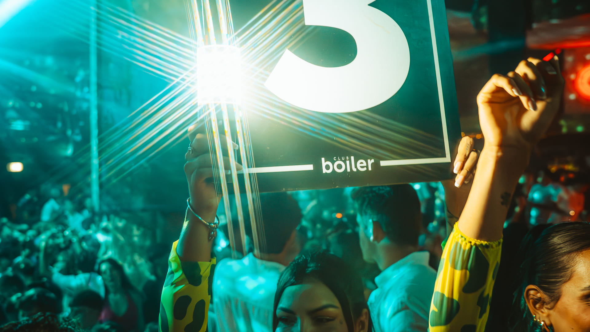 Club Boiler party scene with a glowing number 3 sign held up in a crowd, bathed in vibrant teal light effects.