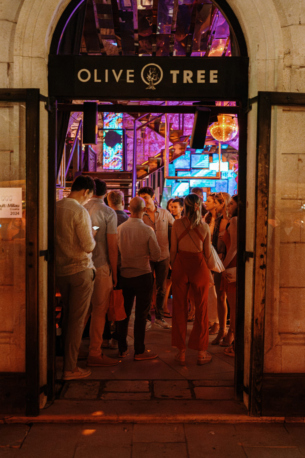 A stylish group of people gathers just inside the entrance of "Olive Tree," a modern nightlife venue. The scene is framed by an arched stone doorway with the venue’s sign above. Inside, colorful LED lighting and mirrors reflect off walls and stairs, creating a vivid, upscale party atmosphere.