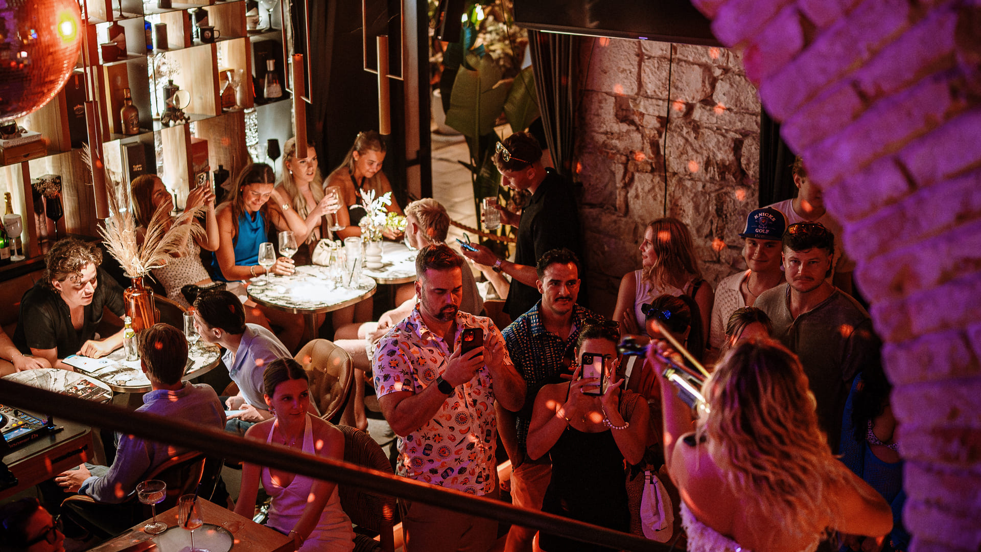 Guests enjoy a lively evening inside a stylish venue with dim lighting, seated at tables with drinks and food, while others stand around recording a performer with their phones. A woman with long blonde hair plays a trumpet in the foreground under red disco lighting.