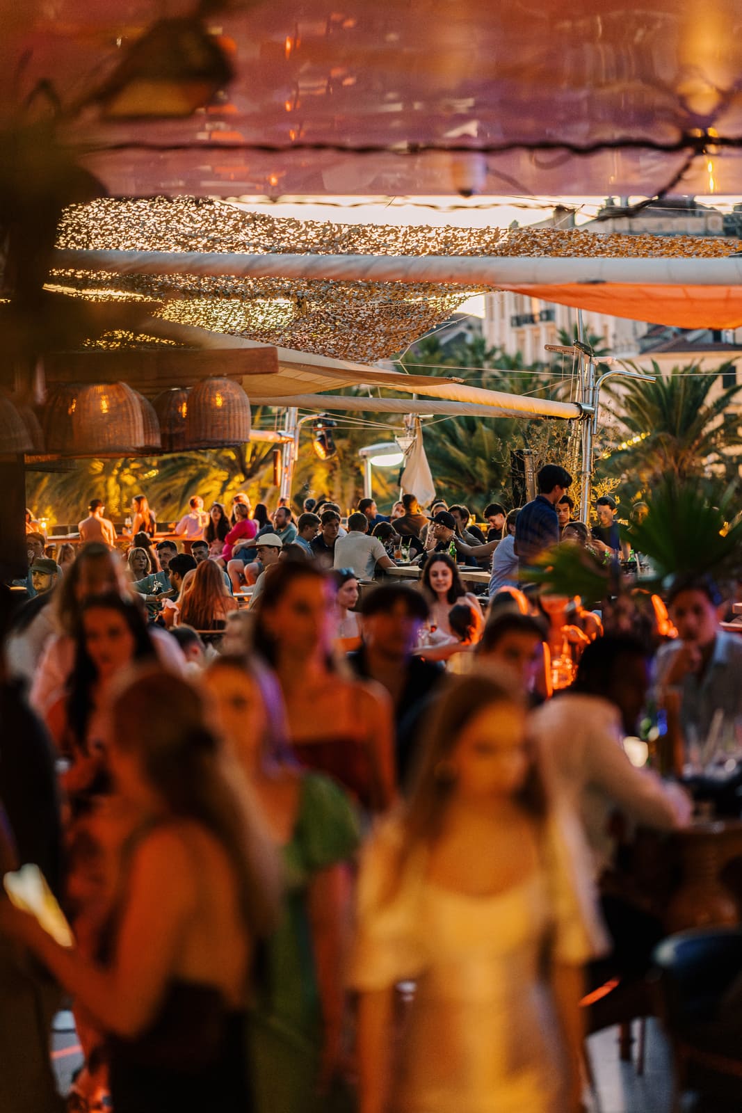 A large crowd fills a chic rooftop bar at sunset, with palm trees and city buildings in the background. The space is shaded by stylish mesh canopies and ambient lighting. In the foreground, elegantly dressed people mingle, while the middle ground shows seated guests enjoying drinks and conversation under a warm golden glow.