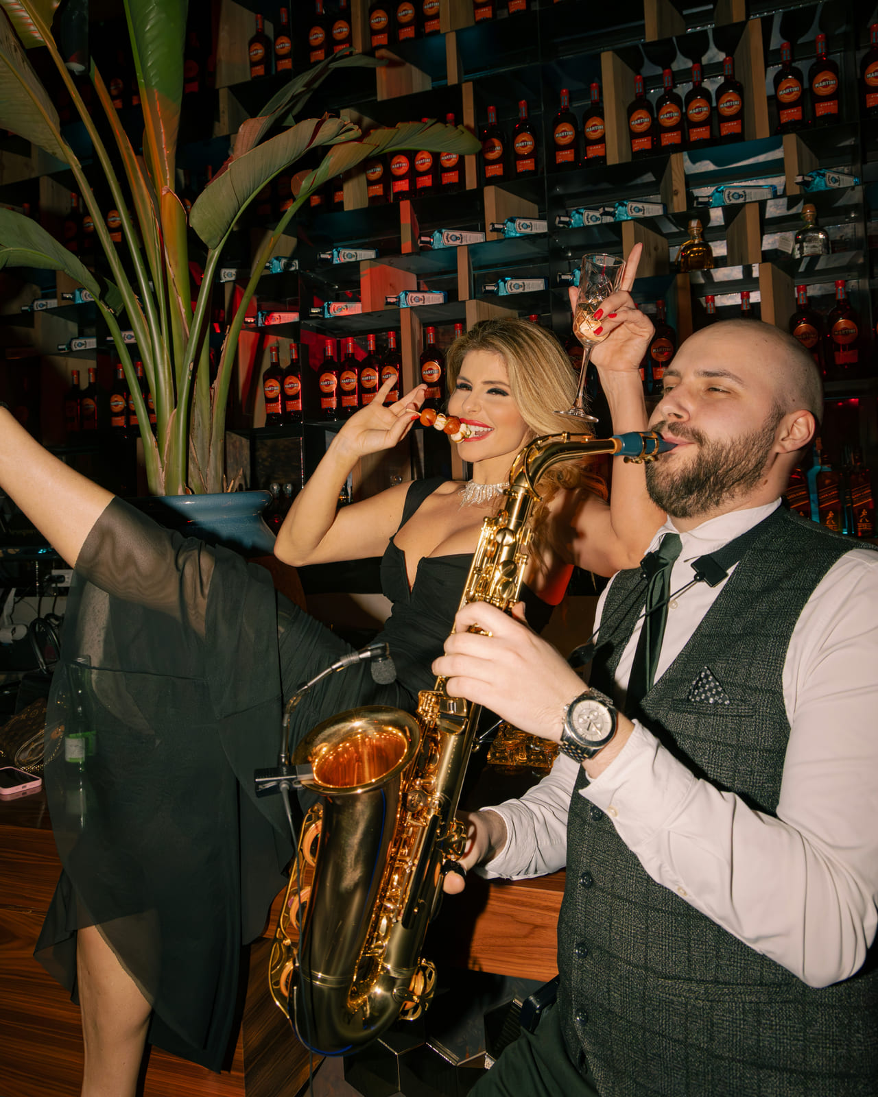 A glamorous woman in a black dress poses playfully with a skewer of fruit while seated next to a saxophonist in formal attire. The background is filled with backlit liquor bottles and tropical plants, suggesting a high-energy, upscale cocktail lounge. Both performers are smiling and appear mid-performance, embodying the venue’s festive atmosphere.