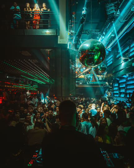 A crowded nightclub scene with a DJ performing on stage, viewed from behind, with green and blue lighting, large disco balls overhead, and a lively audience dancing and enjoying the music.