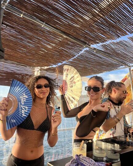 Two stylish women enjoying a rooftop daytime party near the water, holding decorative handheld fans and smiling in front of a DJ booth, with the ocean and sky visible behind them.